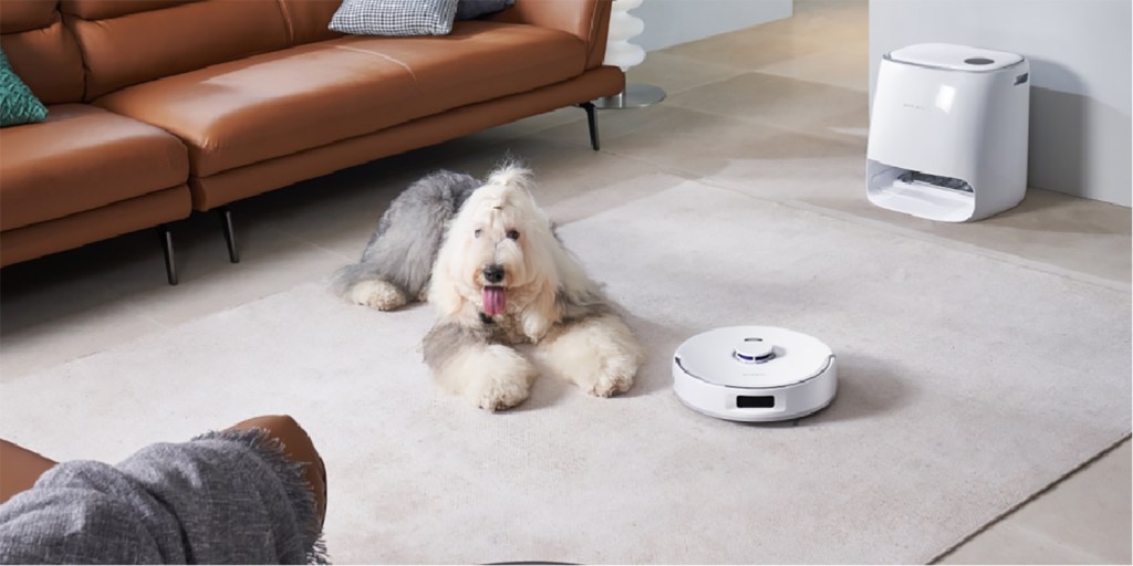 A dog lying on the floor next to a robot vacuum cleaner