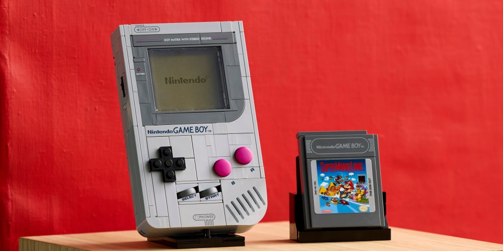 A grey handheld video game console next to a grey handheld video game cartridge. LEGO October 2025