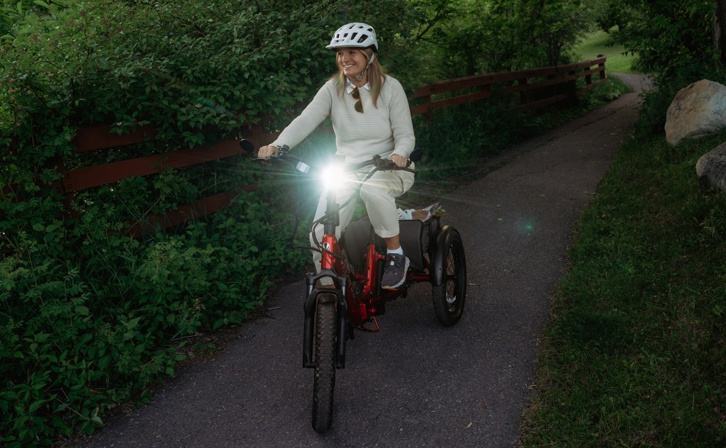 A woman riding Velotric's Triker electric tricycle with headlight on down paved path