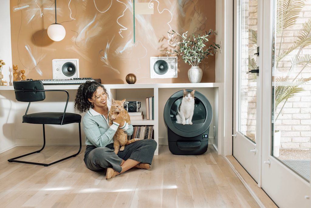 A woman sitting on the floor with two cats in front of a Whisker Litter-Robot EVO