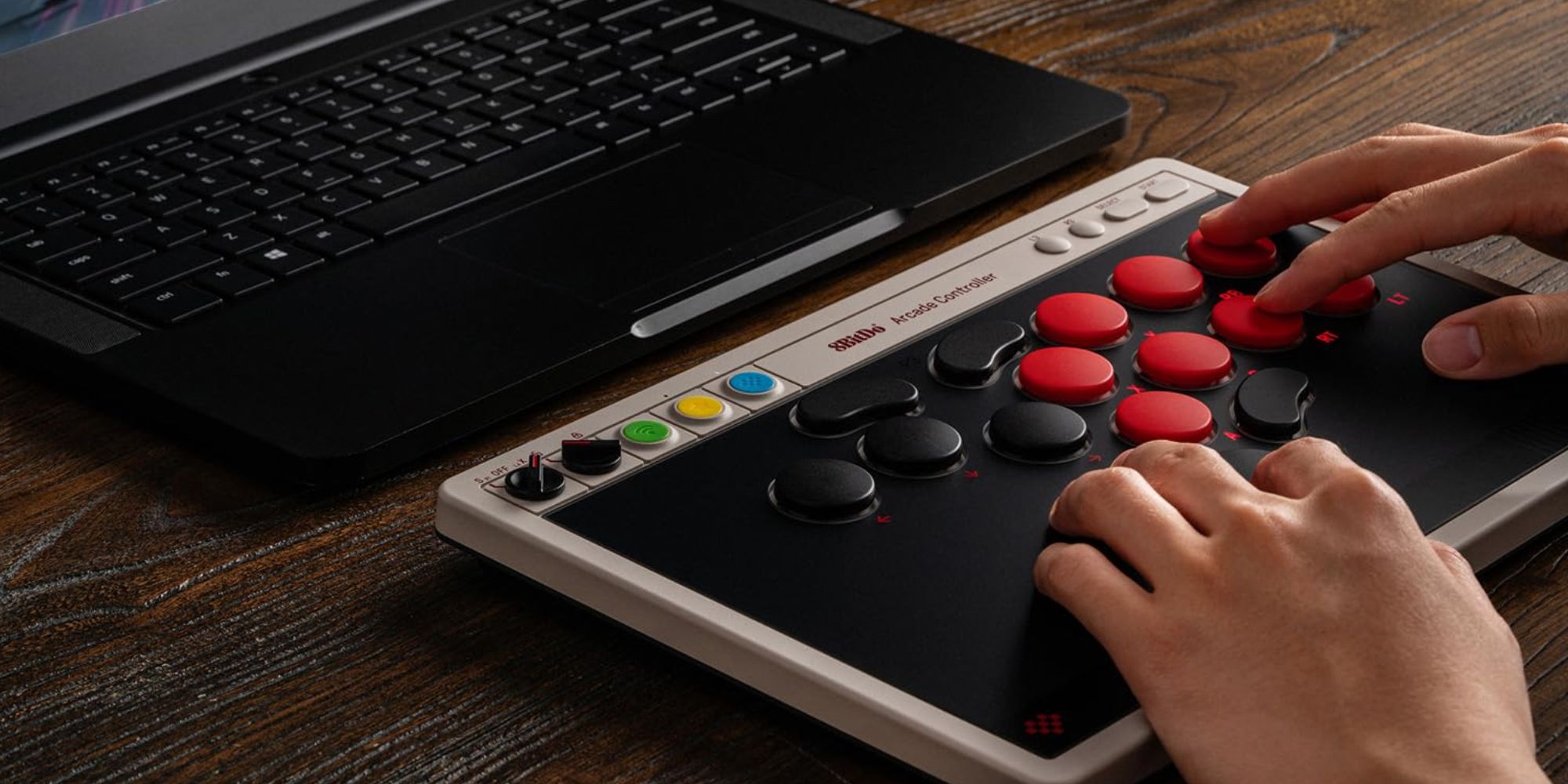 A hand on a black and red controller next to a laptop